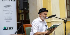 Zdjęcie - na pierwszym planie starszy mężczyzna w czarnym kapeluszu trzyma książkę i czyta jej fragment do mikrofonu. Ma bujną, siwą brodę, białą koszulę i ciemne spodnie. W tle baner TSKŻ i budynek muzeum etnograficznego we Wrocławiu.