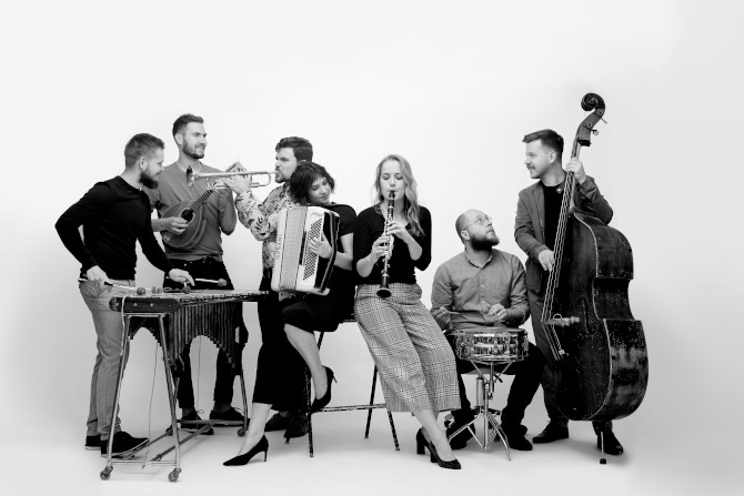 Zespół Warszawskiej Orkiestry Sentymentalnej stoi z instrumentami. Zdjęcie jest czarno-białe, muzycy pozują na jasnym tle. Na zdjęciu jest pięciu mężczyzn i dwie kobiety. 